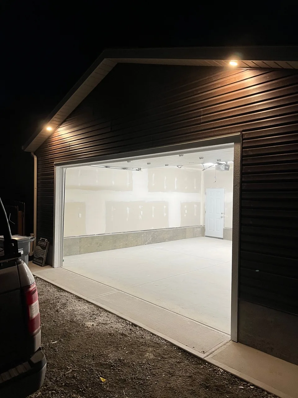 A dark-sided garage at night with the door open, drywalled inside and soffit lights on