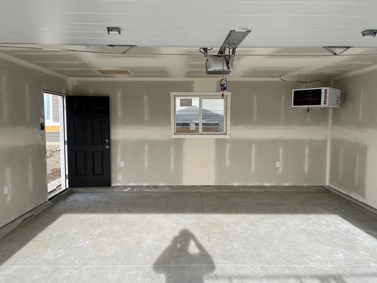 A drywalled garage interior with a black man door, a window and a ceiling-hung unit heater, by Alliance Renovations