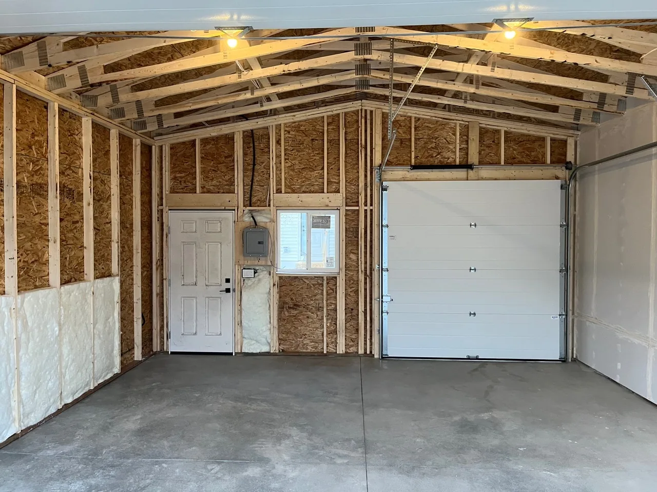 A garage interior with spray-foam insulation, a man door, an electrical panel and a second overhead door